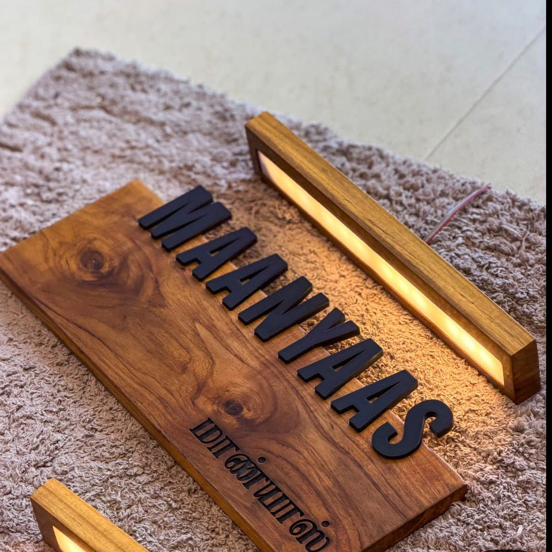 Custom wooden name sign with the name 'MAANYAAS' in raised black letters and a smaller engraved line below, mounted on a rectangular wood plaque. Two slim LED light bars are attached to the sign, casting warm light.